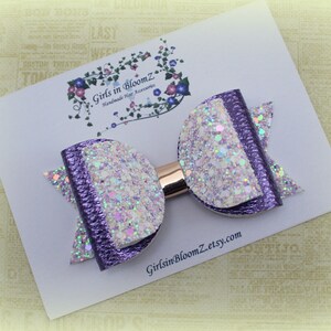 May include: A handmade hair accessory, a purple and white glitter bow, is displayed on a white card. The bow features a metallic gold center and is made of layered materials. The card has the text "Girls in BloomZ Handmade Hair Accessories" and the website address.