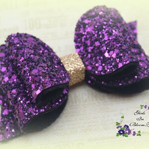 May include: A purple glitter hair bow with a gold center. The bow is made of two layers of glitter fabric and has a black velvet backing.