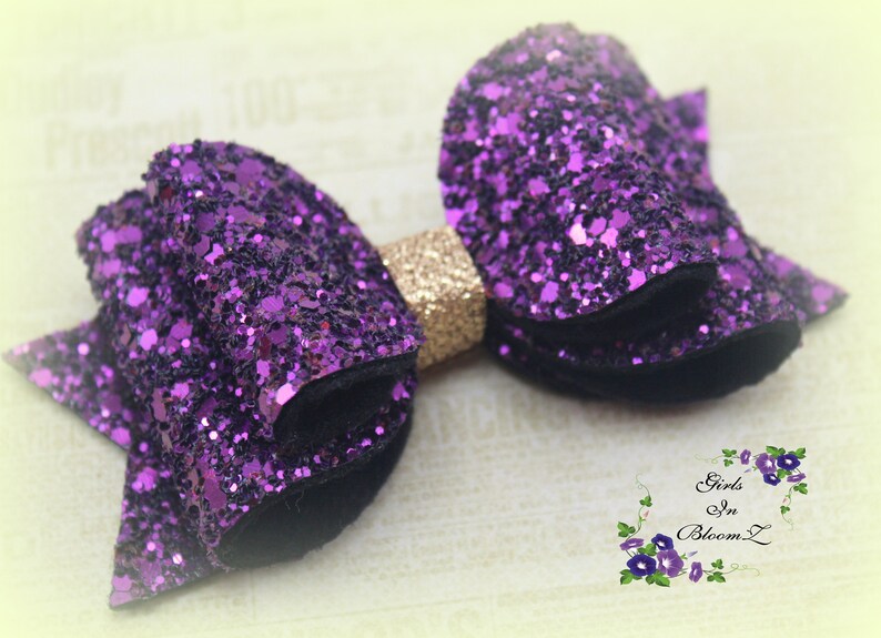 May include: A purple glitter hair bow with a gold center. The bow is made of two layers of glitter fabric and has a black velvet backing.