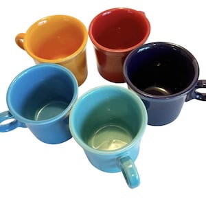 Op de afbeelding: Een set van vijf keramische mokken in verschillende effen kleuren, waaronder oranje, rood, donkerblauw, lichtblauw en turquoise. Elke mok heeft een gladde, glanzende afwerking, een afgerond handvat aan de zijkant en een brede ronde opening.