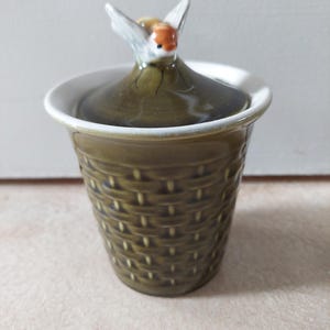 May include: A small olive green ceramic jar with a textured basket-weave pattern on the exterior. The lid features a sculpted, hand-painted bee with white wings and an orange head, serving as a handle for the removable lid of the container.