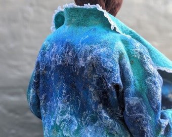 Op de afbeelding: Een vilten jas in blauw- en tinten, met een witte kraag met ruches. Het oppervlak van de jas heeft een getextureerde, gemarmerde uitstraling, met lichtere blauwe en witte accenten. Het ontwerp doet denken aan water of golven. De jas is een uniek draagbaar kunstwerk.