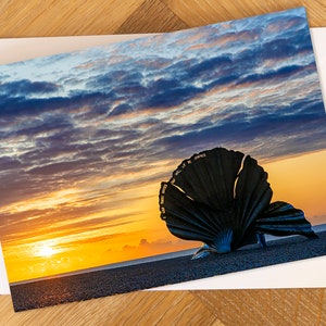 May include: A large, black metal sculpture of a scallop shell on a beach at sunrise. The sky is a vibrant orange and yellow, with clouds in shades of blue and purple. The sculpture is inscribed with the words "I Will Be Your Friend" in gold lettering.