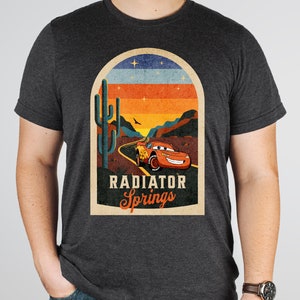 May include: A dark grey t-shirt featuring a vintage-style graphic of a red racing car driving through a desert landscape. The graphic includes a cactus, a sunset, and the text "Radiator Springs".