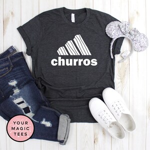 May include: A dark gray t-shirt with a white graphic of a stylized Adidas logo with the word "churro" underneath. The shirt is laying on a white wooden surface with a pair of white sneakers, a pair of blue jeans, and a silver headband with mouse ears.