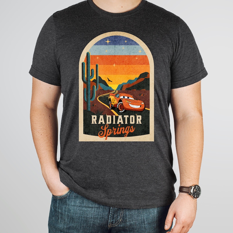 May include: A dark grey t-shirt featuring a vintage-style graphic of a red racing car driving through a desert landscape. The graphic includes a cactus, a sunset, and the text "Radiator Springs".