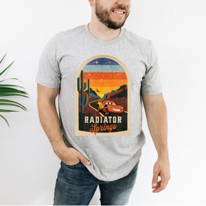 May include: A grey t-shirt featuring a vintage-style graphic of a red racing car driving through a desert landscape with a cactus and the text "Radiator Springs".