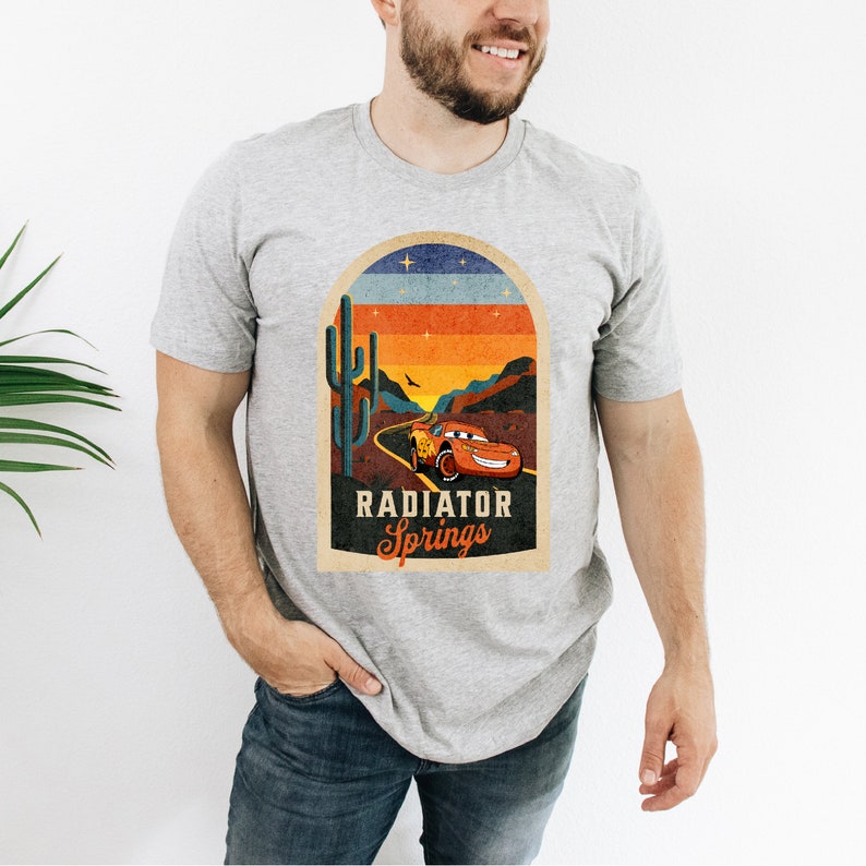 May include: A grey t-shirt featuring a vintage-style graphic of a red racing car driving through a desert landscape with a cactus and the text "Radiator Springs".