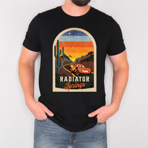 May include: A black t-shirt featuring a vintage-style graphic of a red racing car driving through a desert landscape with a cactus and the text "Radiator Springs".