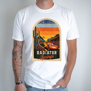 May include: White t-shirt featuring a vintage-style graphic of a red racing car driving through a desert landscape with a cactus and the text "Radiator Springs".