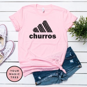 May include: Pink t-shirt with a black graphic of a stylized Adidas logo with the word "churro" underneath. The shirt is laying on a white wooden surface with a pair of white sneakers and a pair of blue denim shorts.