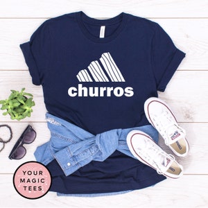 May include: A navy blue t-shirt with a white graphic of a stylized Adidas logo with the word "churro" underneath. The shirt is folded and laid on a wooden surface with a pair of white Converse sneakers and a denim jacket.