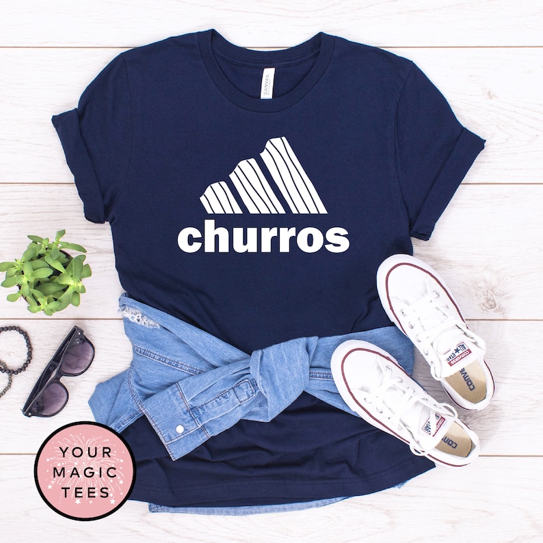 May include: A navy blue t-shirt with a white graphic of a stylized Adidas logo with the word "churro" underneath. The shirt is folded and laid on a wooden surface with a pair of white Converse sneakers and a denim jacket.