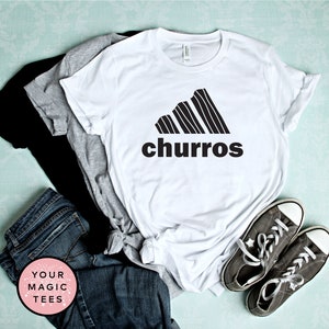 May include: White t-shirt with a black graphic of a churro-shaped Adidas logo and the word "churro" underneath. The shirt is folded and laid on a blue surface with a pair of gray sneakers and a pair of jeans.