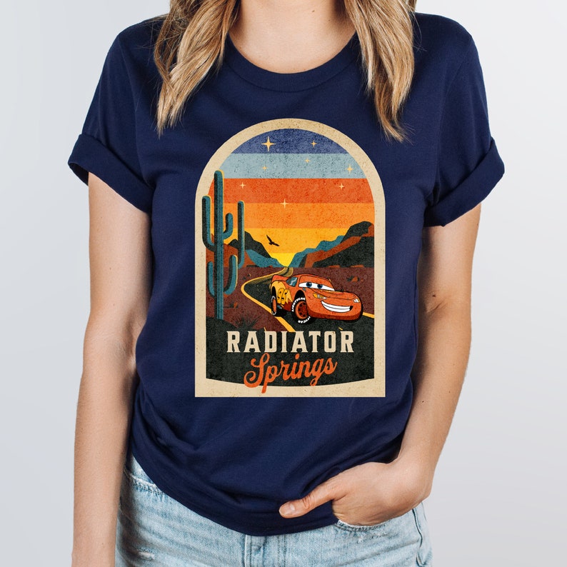 May include: A navy blue t-shirt featuring a vintage-style graphic of a red racing car driving through a desert landscape with a cactus and the text "Radiator Springs".