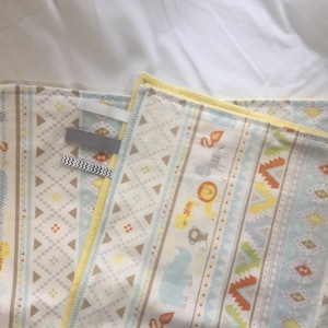 K&ouml;nnte beinhalten: Eine gelbe und blaue Babydecke mit geometrischem Muster und Tierillustrationen. Die Decke hat einen wei&szlig;en Hintergrund mit einem hellblauen, gelben und orangen geometrischen Muster. Die Tiere umfassen einen L&ouml;wen, einen Elefanten, einen Affen und ein Zebra.