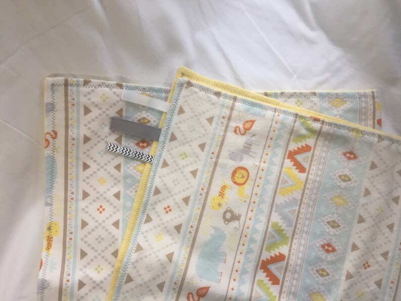 K&ouml;nnte beinhalten: Eine gelbe und blaue Babydecke mit geometrischem Muster und Tierillustrationen. Die Decke hat einen wei&szlig;en Hintergrund mit einem hellblauen, gelben und orangen geometrischen Muster. Die Tiere umfassen einen L&ouml;wen, einen Elefanten, einen Affen und ein Zebra.