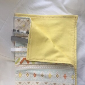 K&ouml;nnte beinhalten: Eine gelbe Stoffdecke f&uuml;r Babys mit einem geometrischen Muster in Blau-, Braun- und Wei&szlig;t&ouml;nen. Die Decke ist in der H&auml;lfte gefaltet und hat ein wei&szlig;es Etikett.