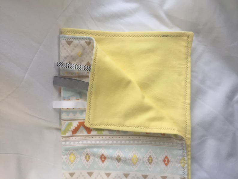 K&ouml;nnte beinhalten: Eine gelbe Stoffdecke f&uuml;r Babys mit einem geometrischen Muster in Blau-, Braun- und Wei&szlig;t&ouml;nen. Die Decke ist in der H&auml;lfte gefaltet und hat ein wei&szlig;es Etikett.