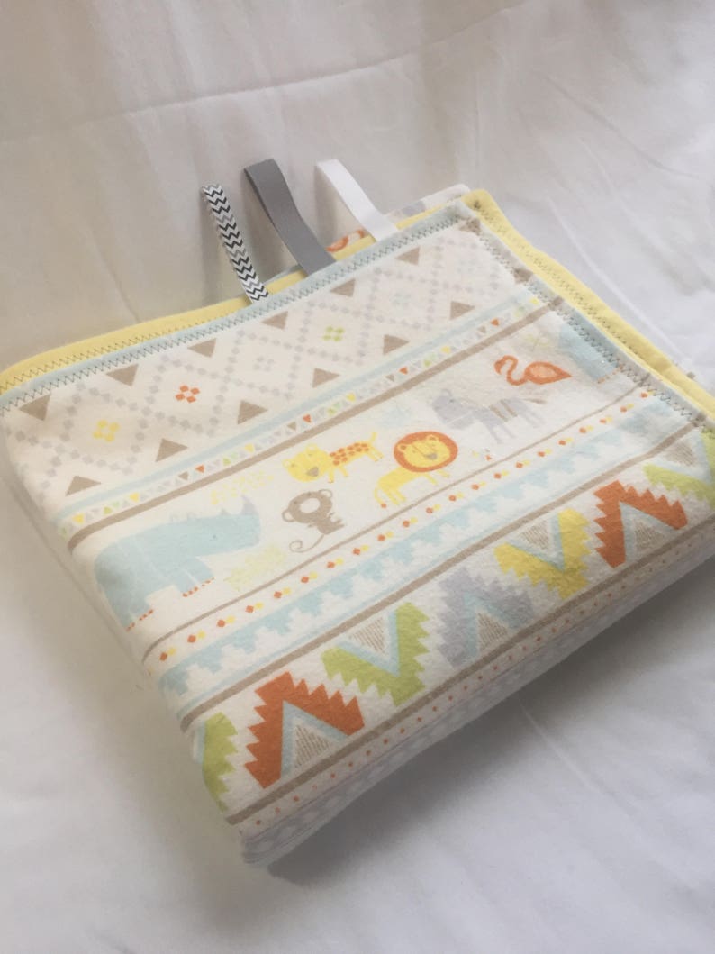 K&ouml;nnte beinhalten: Eine wei&szlig;e und gelbe Babydecke mit geometrischem Muster und Tierillustrationen. Die Decke hat zwei Stoffschlaufen zum Aufh&auml;ngen.