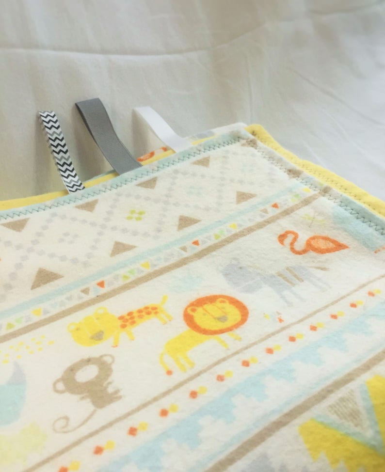 K&ouml;nnte beinhalten: Eine wei&szlig;e und gelbe Babydecke mit einem bunten Tierdruck. Die Decke hat ein hellblaues, gelbes und braunes geometrisches Muster. Die Tiere sind ein Affe, ein L&ouml;we, eine Giraffe und ein Flamingo.