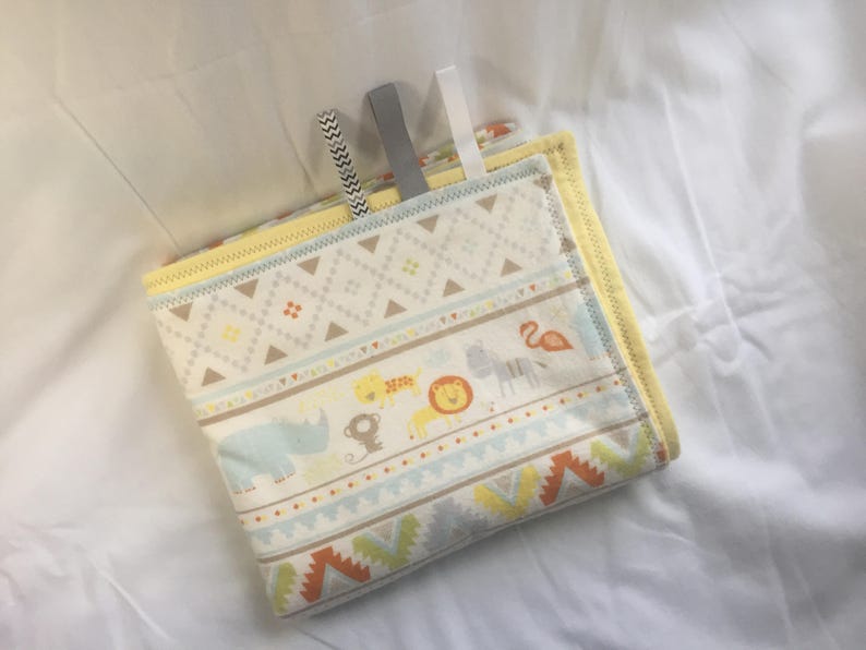 K&ouml;nnte beinhalten: Eine gelbe und wei&szlig;e Babydecke mit geometrischem Muster und Tierillustrationen. Die Decke hat einen gelben Rand und ist in der H&auml;lfte gefaltet.