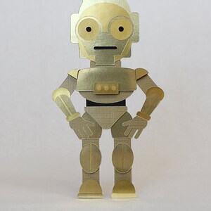 May include: A gold robot figurine with a black belt and three round buttons. The robot has a small antenna on its head and is standing with its hands on its hips.