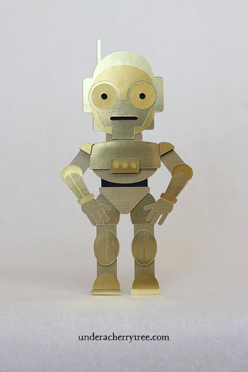 May include: A gold robot figurine with a black belt and three round buttons. The robot has a small antenna on its head and is standing with its hands on its hips.