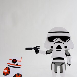 May include: A white and gray paper cutout of a Stormtrooper from Star Wars holding a black blaster and aiming it at a white and orange paper cutout of BB-8.