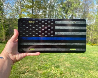 May include: A black and white American flag with a thin blue line running horizontally across the middle of the flag. The flag is printed on a metal license plate.