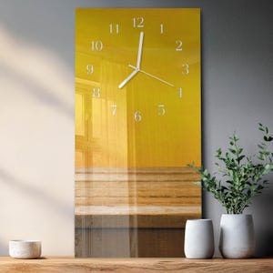 May include: A rectangular wall clock with a yellow background and white clock hands and numbers. The clock face features a blurred image of a room with a window. The clock is displayed on a wooden surface, with a grey wall and plants in white pots in the background.
