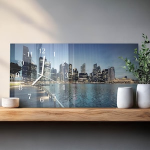 May include: A rectangular wall clock with a cityscape design. The clock face features a nighttime view of a city skyline with tall buildings and a body of water. The clock hands are white, and the numbers are visible.