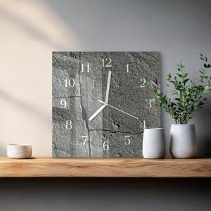 May include: Square wall clock with a grey stone texture. The clock face has white numerals and hands. The clock is displayed on a wooden shelf with white vases and a potted plant.