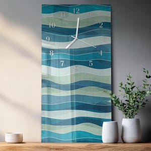 May include: A rectangular wall clock with a wavy pattern in shades of blue and green. The clock face has silver-colored hands and numerals. The clock is displayed on a wooden surface, with white vases and greenery in the background.