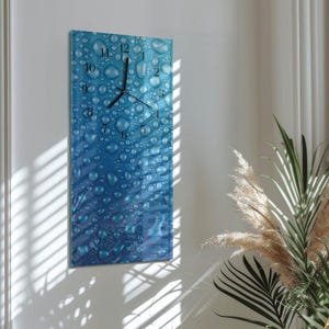 May include: A rectangular wall clock with a blue water droplet design. The clock face features black hands and numerals. The clock is mounted on a white wall, with natural light and plants in the background.