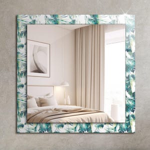 Puede incluir: Un espejo rectangular con fondo blanco y un borde de hojas tropicales verdes. El espejo refleja un dormitorio con una cama, una lámpara y una ventana.