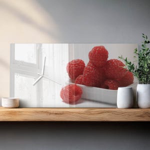 May include: Rectangular wall clock with a white face, window design, and white hands. A bowl of fresh red raspberries is on the clock face. The clock is displayed on a wooden shelf.