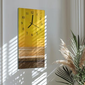 May include: A rectangular wall clock with a yellow and brown gradient design. The clock face features black numerals and hands. The clock is mounted on a white wall, with natural light and plants in the background.