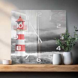 Könnte beinhalten: Eine quadratische Wanduhr mit einer schwarz-weiß Fotografie einer Küstenlandschaft und einem rot-weiß gestreiften Leuchtturm. Das Zifferblatt zeigt große weiße Ziffern von 1 bis 12 sowie zwei dünne silberne Zeiger in der Mitte des Bildes.