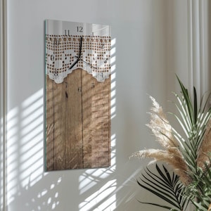 Op de afbeelding: Een rechthoekige wandklok met een houten plankontwerp en een witte kanten overlay. De wijzerplaat heeft zwarte cijfers en wijzers. De klok is aan een witte muur bevestigd.