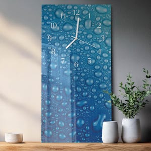Puede incluir: Un reloj de pared rectangular con un fondo degradado azul que presenta un patrón de gotas de agua. Las manecillas y los números blancos indican la hora. El reloj se exhibe sobre una superficie de madera, con plantas y jarrones decorativos cerca.