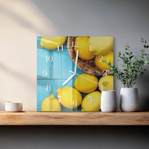 May include: A square wall clock featuring a printed image of bright yellow lemons in a woven basket against a light blue wooden background. The clock face displays white numbers from 1 to 12 and two white hands, indicating the current time clearly.