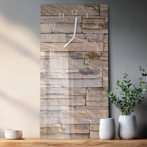 May include: A rectangular wall clock with a stone-like texture background. The clock face features white numerals and hands, indicating the time. The clock is mounted on a gray wall, with decorative plants in white pots on a wooden surface.