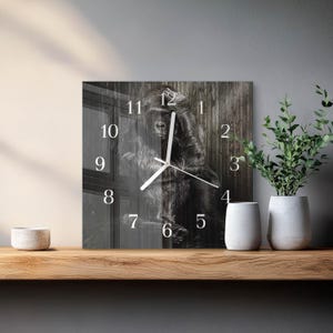 May include: Square wall clock featuring a black and white image of a gorilla. The clock face has white numerals and hands. The background shows a wooden texture. The clock is displayed on a wooden shelf with decorative items.