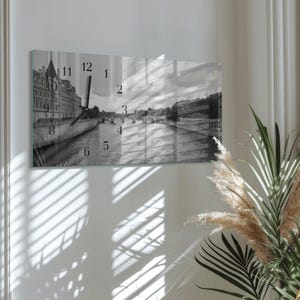 Könnte beinhalten: Rechteckige Wanduhr in Schwarzweiß mit einer europäischen Stadtlandschaft mit Fluss und Brücken. Das Zifferblatt hat schwarze Ziffern und Zeiger. Die Uhr ist an einer weißen Wand befestigt.