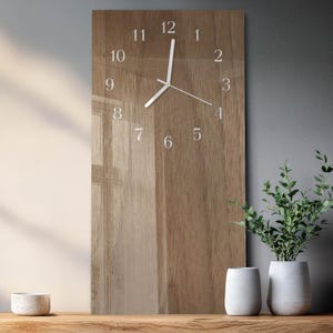 Op de afbeelding: Een rechthoekige wandklok met een houtnerfpatroon. De wijzerplaat heeft witte cijfers en wijzers. De klok wordt weergegeven op een houten oppervlak, met witte vazen en groen rechts en een kleine kom links.