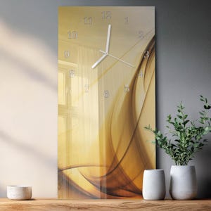 May include: A rectangular wall clock with a gold and brown abstract design. The clock face features silver numerals and hands. The clock is displayed on a wooden surface with decorative vases and plants.