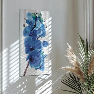 May include: A rectangular wall clock featuring a vibrant blue orchid design. The clock face displays black numerals and hands against the floral backdrop. The clock is mounted on a white wall, with natural light and shadows.