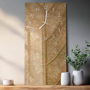 May include: A rectangular wall clock with a detailed leaf design in shades of brown and tan. The clock face has white numerals and hands. The clock is mounted on a grey wall, with decorative plants and vases on a wooden surface.