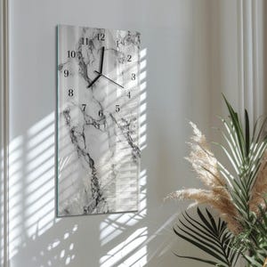 Può includere: Un orologio da parete rettangolare con un design marmorizzato. Il quadrante dell'orologio presenta numeri e lancette nere su uno sfondo bianco e grigio. L'orologio è montato su una parete bianca.